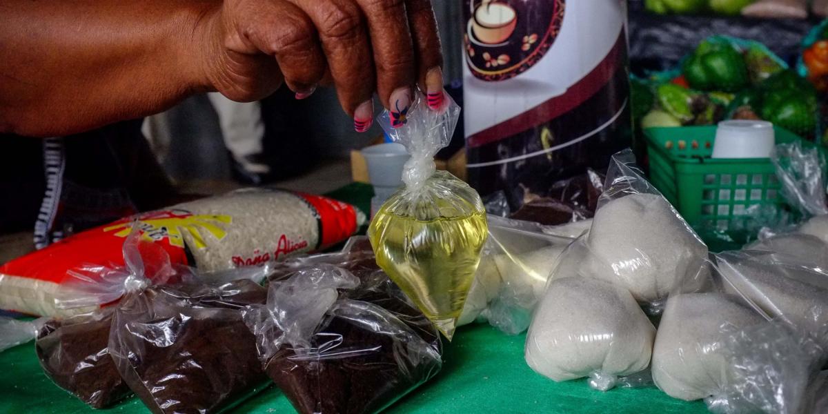 La imagen muestra a una vendedora informal mientras sostiene una bolsa de aceite vegetal, que ofrece a la venta junto a azúcar y café en un puesto en una calle de Caracas (Venezuela).