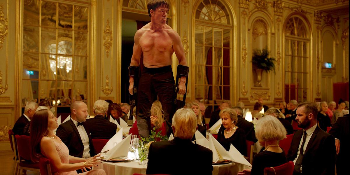 El actor Terry Notary (sobre la mesa) interpreta a Oleg, quien realiza un ‘performance’ imitando a un mono en medio de una cena.