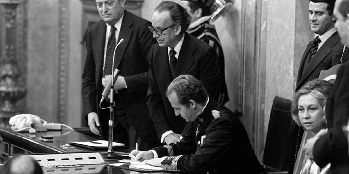 En el congreso de los Diputados, el rey Juan Carlos I firma la nueva Constitución de los españoles. A su lado, la reina Sofía.