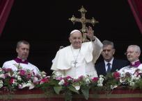 Se ve al Papa Francisco, acompañado de otros sacerdotes, desde el balcón. Él est´pa levantando la mano, saludando a los presentes. Detrás de él está un crucifijo.