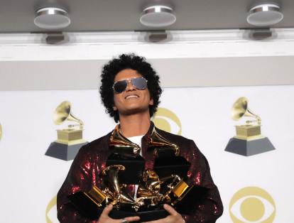 16-	Bruno Mars, en medio de su brillante chaqueta y frente a la pared de los Grammy, junta con fuerza sus brazos, para poder llevar los premios que ganó gracias a su recorrido musical exitoso.