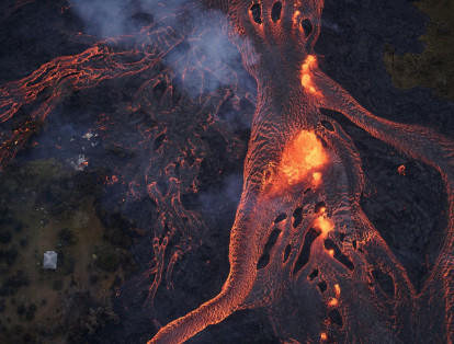 Un gran río de lava a punto de consumir una casa, en la parte derecha de la fotografía, mientras siguen ardiendo los restos de otra casa, en la parte izquierda.