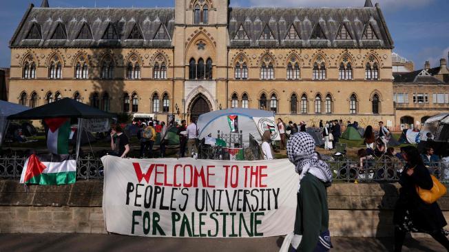 Un transeúnte con la cabeza cubierta con una keffiyeh pasa junto a una pancarta que dice "Bienvenidos a la universidad popular para Palestina" en la Universidad de Oxford, en Oxford, este de Inglaterra, el 7 de mayo de 2024, mientras se ha establecido un campamento pro-palestino en el instalaciones.